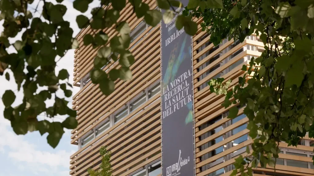 Fachada del edificio del IRB Lleida con una lona vertical sobre investigación, enmarcada por hojas de un árbol, grabado para un vídeo corporativo.